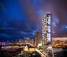 High-rise residential tower featuring a glass facade, recessed balconies, a tiered podium structure, and an illuminated exterior design