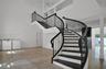 Curved staircase featuring dark wood treads and a black metal railing, complemented by double-height ceilings, stone tile flooring, and a glass-panele