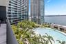 Private balcony with wood-finish flooring, overlooking a large swimming pool, landscaped palm trees, and waterfront horizon