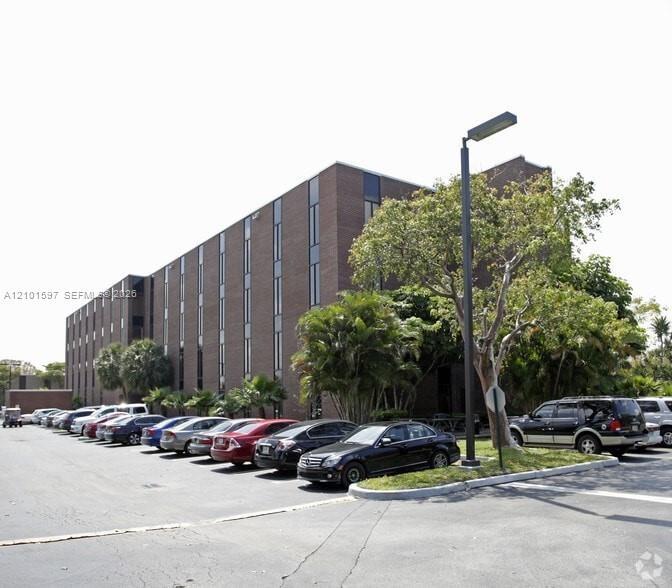 Four-story masonry exterior featuring vertical glass paneling, paved surface parking lot, and mature perimeter landscaping