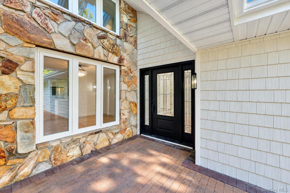 Exterior entrance featuring multi-hued natural stone masonry, white shake siding, brick paver porch, and a black front door with decorative glass pane