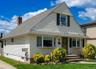 Featuring cream-colored siding, a steeply pitched roof, bay windows, and a brick chimney