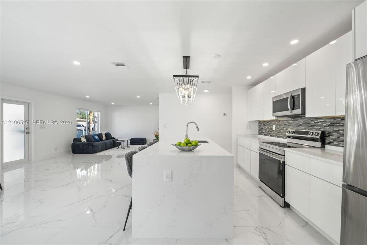 Kitchen featuring white flat-panel cabinetry, a central island with a solid-surface waterfall countertop, stainless steel appliances, a glass-tile bac
