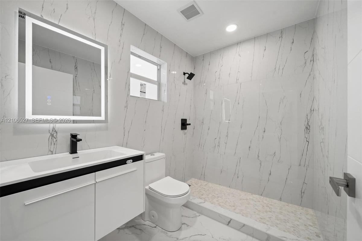 Bathroom featuring full-height stone-finish wall tile, walk-in shower with pebble-stone flooring, integrated LED vanity mirror, white cabinetry with a