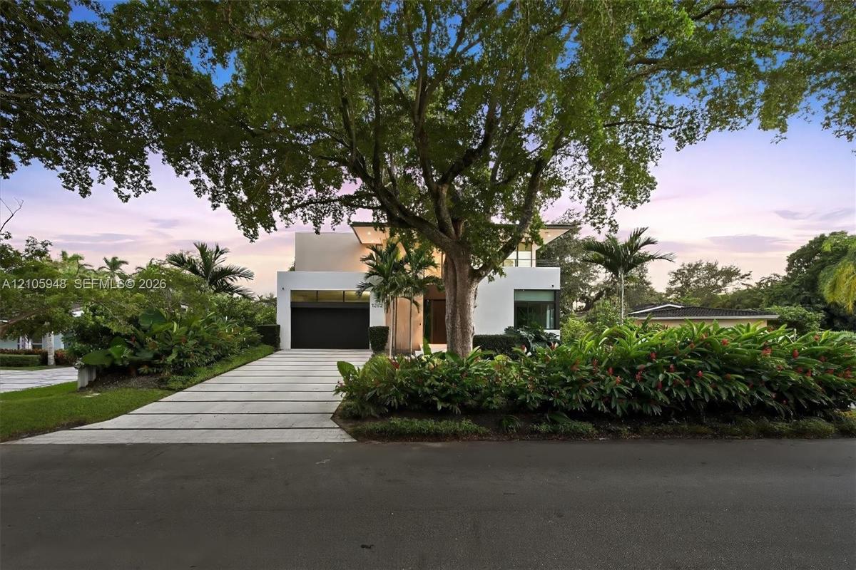 Modern architectural facade featuring a smooth stucco exterior, linear concrete paver driveway, dark-finish multi-car garage door, and lush tropical l