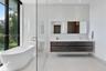 Freestanding oval soaking tub, wall-mounted dark wood-finish dual vanity with integrated sinks, large-format tile flooring, floor-to-ceiling glass-enc
