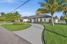Single-story residence featuring a low-slope roof, white exterior cladding, a curved paved driveway, and established tropical landscaping