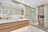 Floating wood-finish double vanity with rectangular integrated sinks, expansive LED-backlit mirror, and glass-enclosed shower featuring a river-stone 
