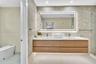 Floating wood-finish vanity with dual integrated sinks, quartz countertop, illuminated LED vanity mirror, full-height stone-finish wall tile, and glas
