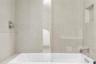 Soaking tub featuring large-format neutral tile surround, integrated wall niche, chrome wall-mounted faucet, and glass partition