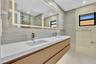 Double vanity featuring a white composite countertop, integrated rectangular sinks, wood-finish cabinetry, a large back-lit mirror, and neutral wall-t