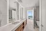 Double vanity featuring light-toned wood cabinetry, large-format tile flooring, integrated wall-mounted mirrors with backlighting, and a glass-enclose
