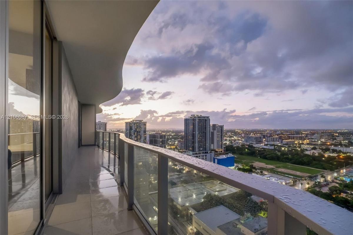 Covered balcony with a curvilinear roofline, glass panel railings, and tile flooring, featuring expansive urban surroundings