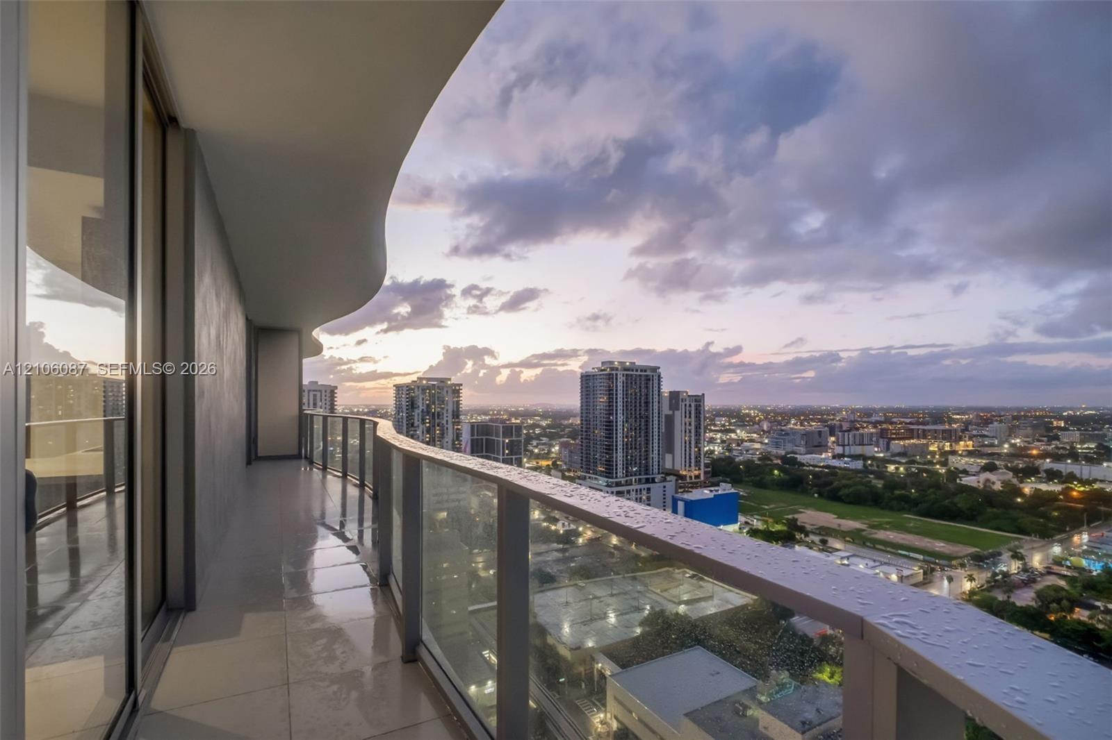 Covered balcony with a curvilinear roofline, glass panel railings, and tile flooring, featuring expansive urban surroundings