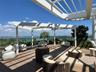 Expansive rooftop terrace with wood-finish decking, white pergola structure, integrated landscaping, and distant city and water scenery