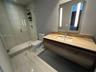 Floating wood-finish vanity with an integrated countertop sink, wall-mounted mirror with integrated lighting, glass-enclosed shower, and large-format 