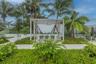 Outdoor patio featuring a white slatted pergola, integrated ceiling fan, light-colored paver flooring, raised garden beds, and mature palm trees
