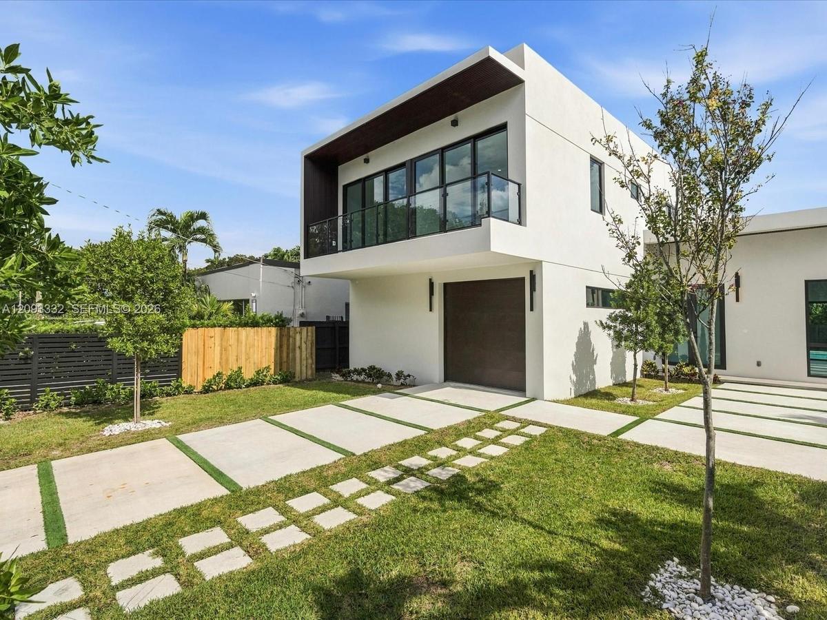Modern stucco exterior featuring a cantilevered balcony, dark-tinted glass railings, multi-panel sliding doors, and a decorative concrete paver drivew