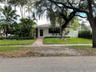 Single-story residence featuring a tile roof, light-colored exterior finish, brick paver walkway, mature landscaping, and white privacy fencing