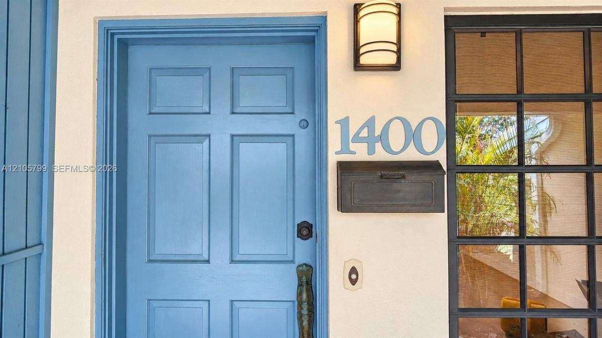 Entryway featuring a blue-painted paneled door, black metal wall-mounted mailbox, brushed metal house numbers, and a dark-framed grid-style sidelight