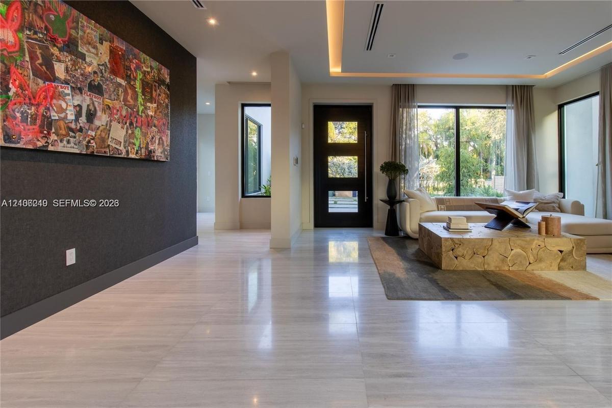 Large-format polished stone flooring, recessed tray ceiling with integrated cove lighting, dark solid-core entry door with glass inserts, and floor-to