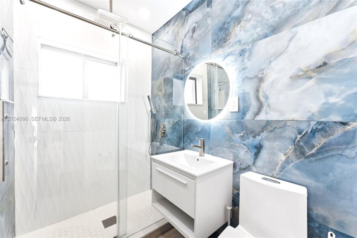 Large-format blue stone-finish wall tile, frameless glass shower enclosure with rain-style showerhead, floating vanity, circular backlit mirror, and w