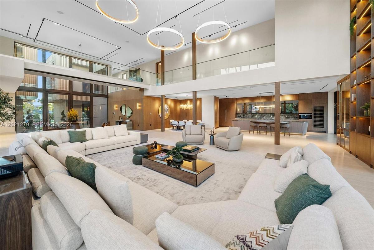 Double-height great room with triple ring chandelier, wraparound sectional seating, light stone floors, open second-floor glass balcony, walnut column
