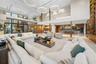 Double-height great room with triple ring chandelier, wraparound sectional seating, light stone floors, open second-floor glass balcony, walnut column