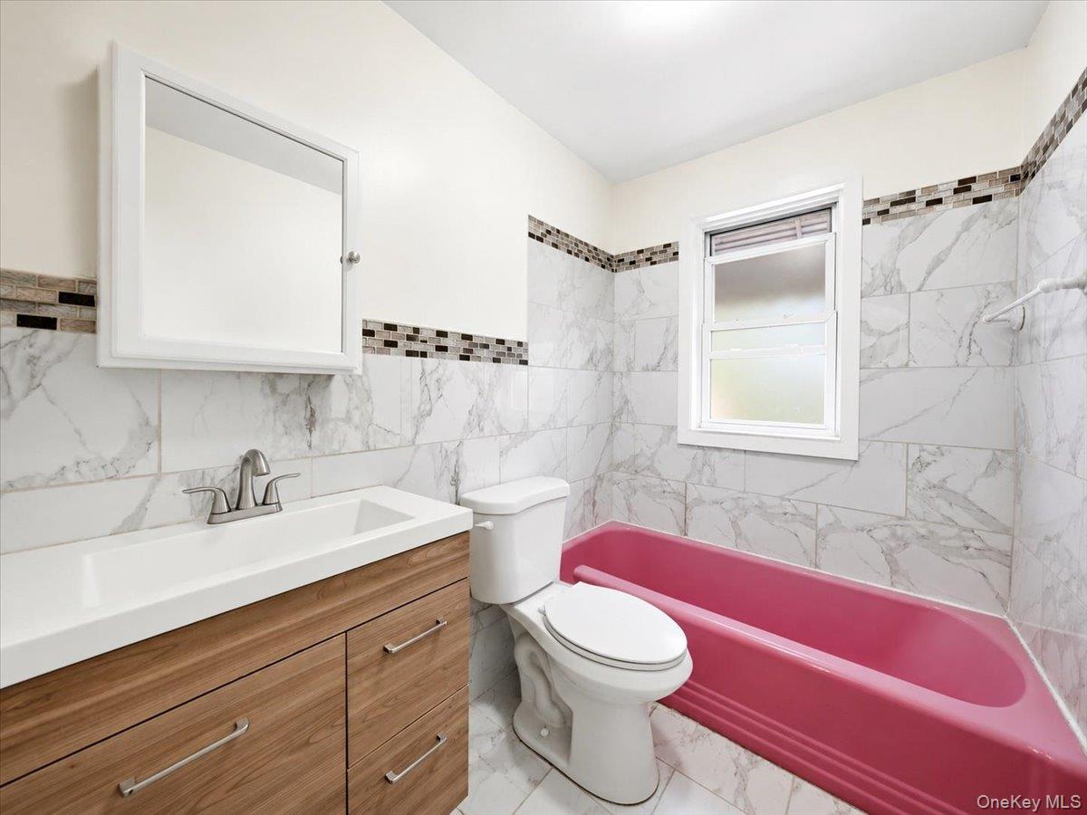 White tiled walls with a decorative mosaic accent, wood-finish vanity, integrated white sink, and a white framed window