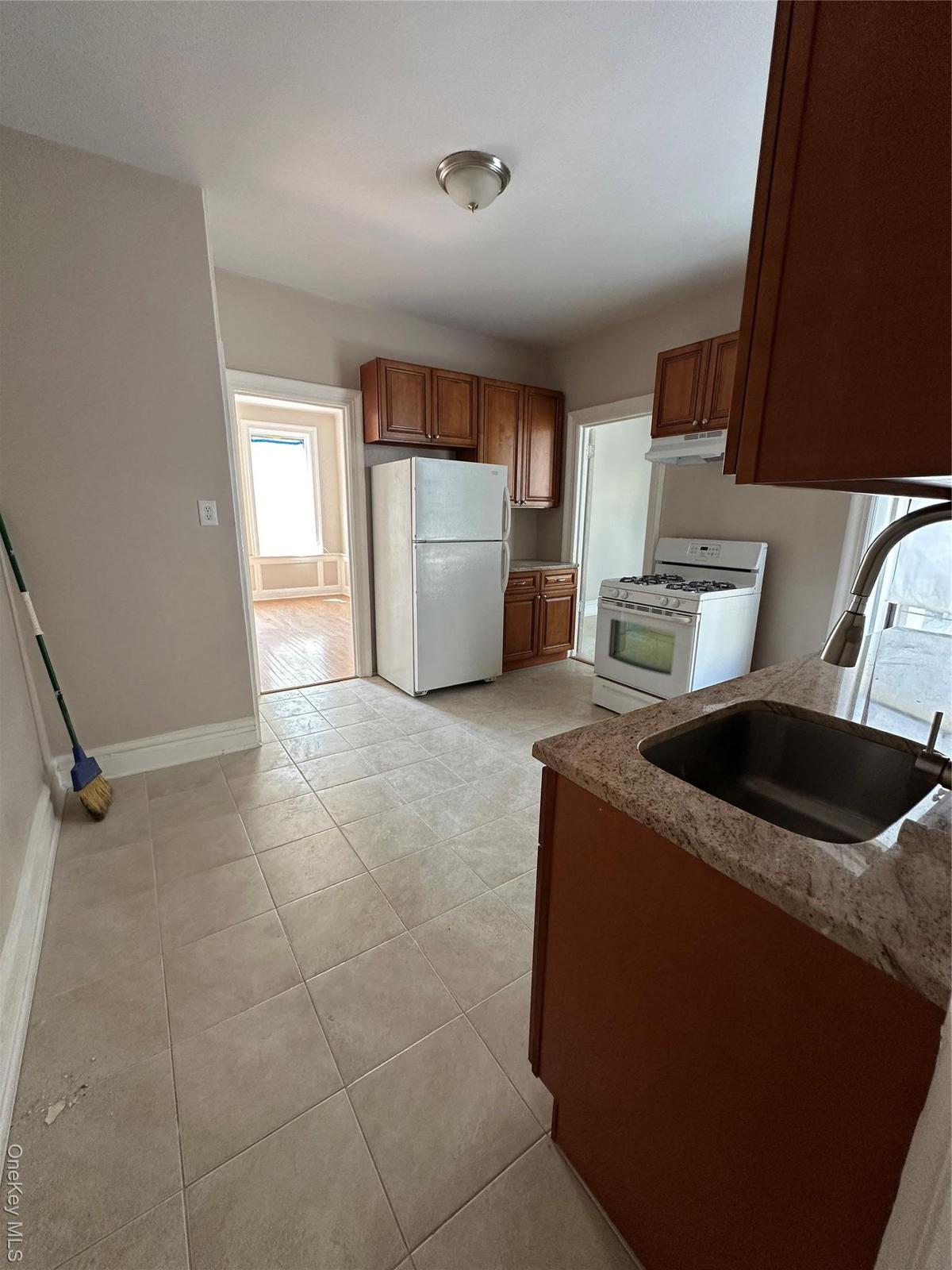 Kitchen with stove, freestanding refrigerator, brown cabinetry, light stone counters, and light tile patterned floors