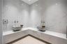 Spa-quality steam room with full-height polished white marble tile walls, corner L-shaped bench, dual ornate carved white vessel sinks, antique brass