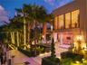 Dusk view of waterfront side yard with string-lit royal palms, manicured hedges, illuminated walkway, dock access, covered loggia with outdoor dining