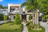 Two-story modern luxury home with stone façade, paver walkway, bronze sculpture, manicured hedges, tropical landscaping, second-level glass balcony, a