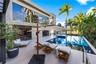 Large-format stone pool deck with teak chaise lounges, cantilever umbrella, woven daybed, dark mosaic tile pool, stone-clad two-story rear facade, flo