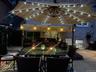 Paved outdoor patio featuring an illuminated umbrella, a built-in bar with a thatched roof accent, and a privacy screen of artificial greenery