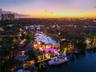 Aerial dusk view of modern waterfront estate with illuminated pool, covered loggia, manicured grounds, private dock with yacht, programmable LED light