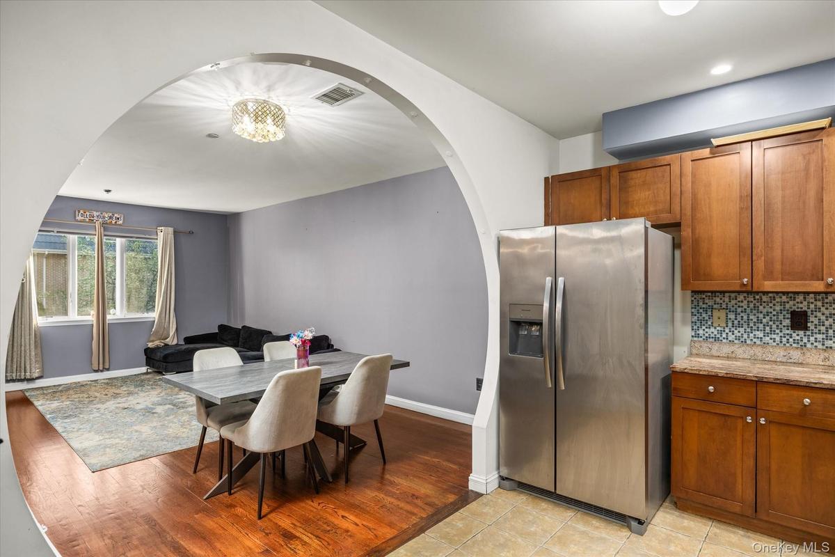 Kitchen and dining area featuring an arched doorway, wood-finish flooring, stainless steel refrigerator, wood cabinetry, and a mosaic tile backsplash