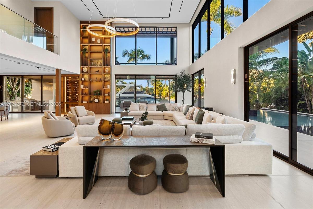 Sun-filled double-height great room with oversized curved sectional, dual ring chandelier, full-height illuminated walnut shelving, wine display, floo