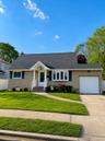 Shingle siding facade with dark blue shutters, covered entry with white railing, attached garage, paved driveway, and established front lawn