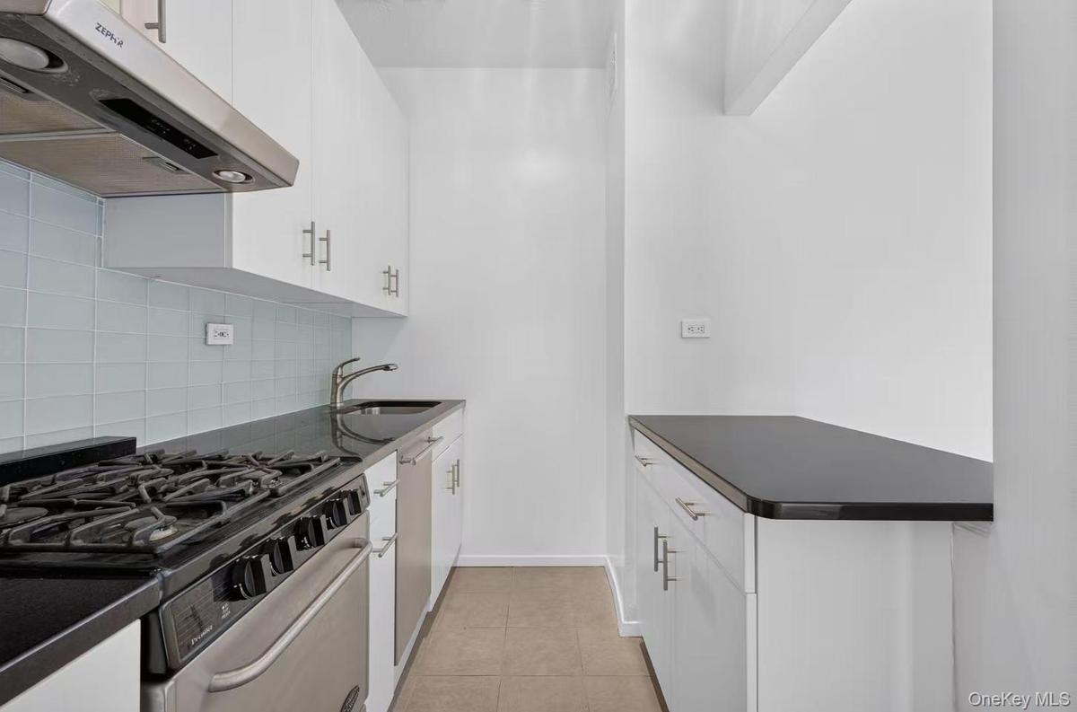 Galley kitchen featuring white cabinetry, dark countertops, a gas range with stainless steel oven, an undermount sink, and a light blue tiled backspla