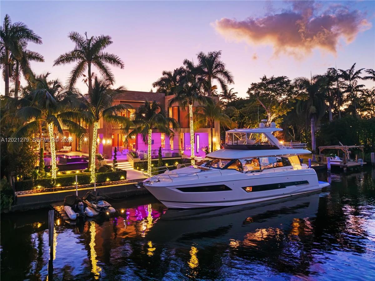 Ground-level dusk view from canal of private deep-water dock with large motor yacht, illuminated seawall, string-lit palms, manicured waterfront lawn,