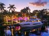 Ground-level dusk view from canal of private deep-water dock with large motor yacht, illuminated seawall, string-lit palms, manicured waterfront lawn,