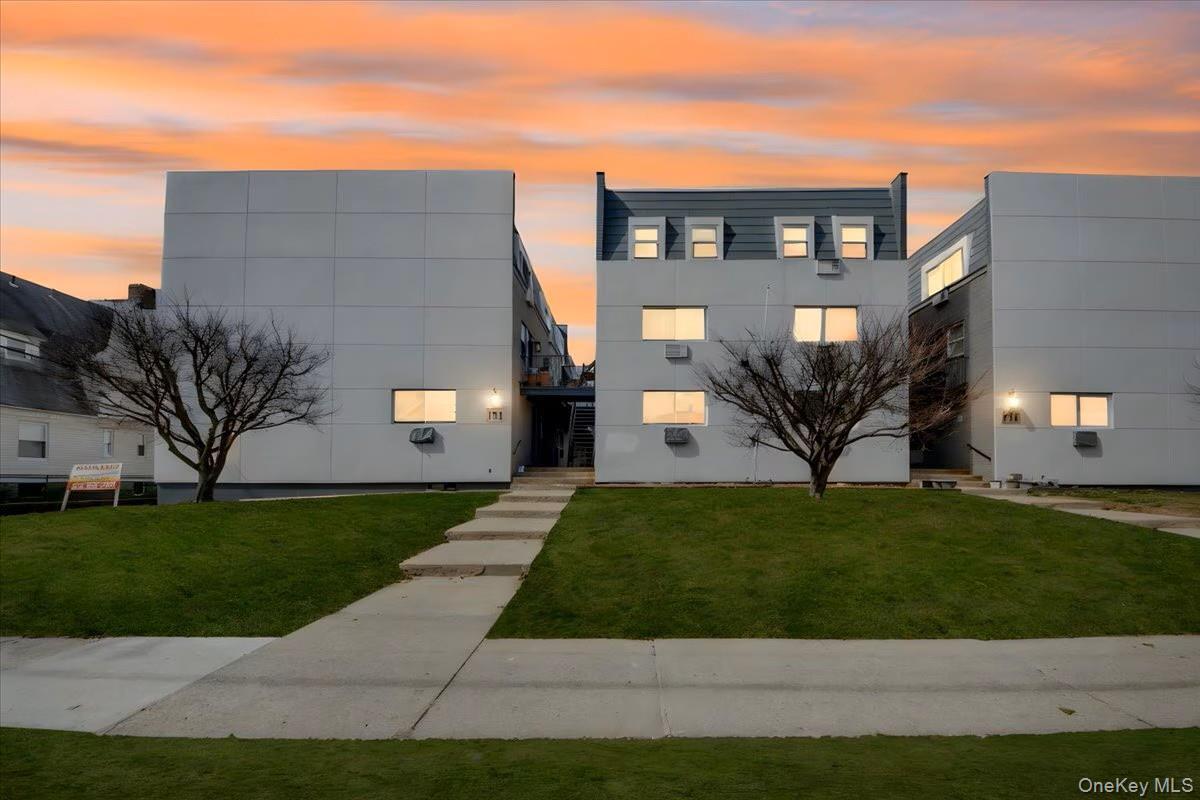 Modern multi-story building featuring a light-colored facade, multiple windows, dormer windows, and a manicured lawn with a concrete pathway