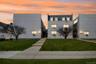 Modern multi-story building featuring a light-colored facade, multiple windows, dormer windows, and a manicured lawn with a concrete pathway