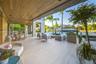 Expansive waterfront ground-level terrace with large-format stone tile, covered loggia with outdoor kitchen and dining, multiple woven lounge seating