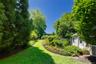 Landscaped grounds featuring a winding grassy path, mature trees, dense shrubbery, a white lattice fence, and a white shed with blue shutters