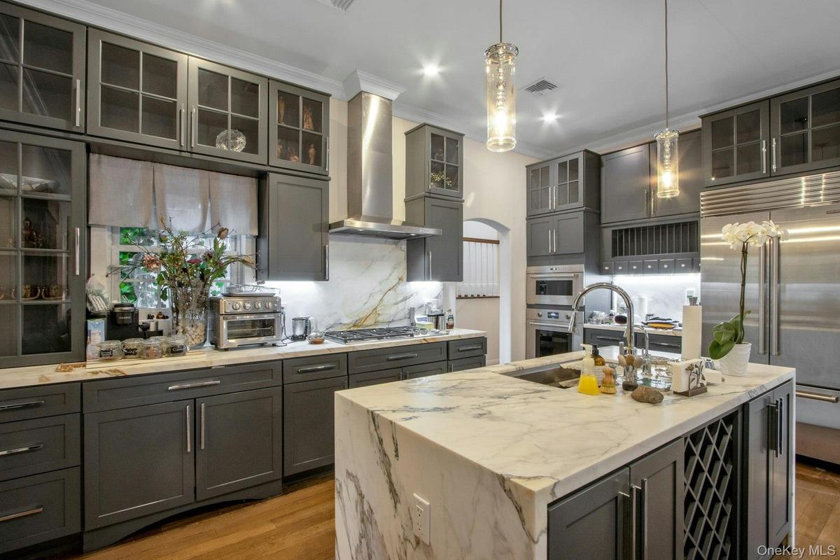 Kitchen featuring wall chimney exhaust hood, stainless steel appliances, light stone counters, an island with sink, and decorative light fixtures