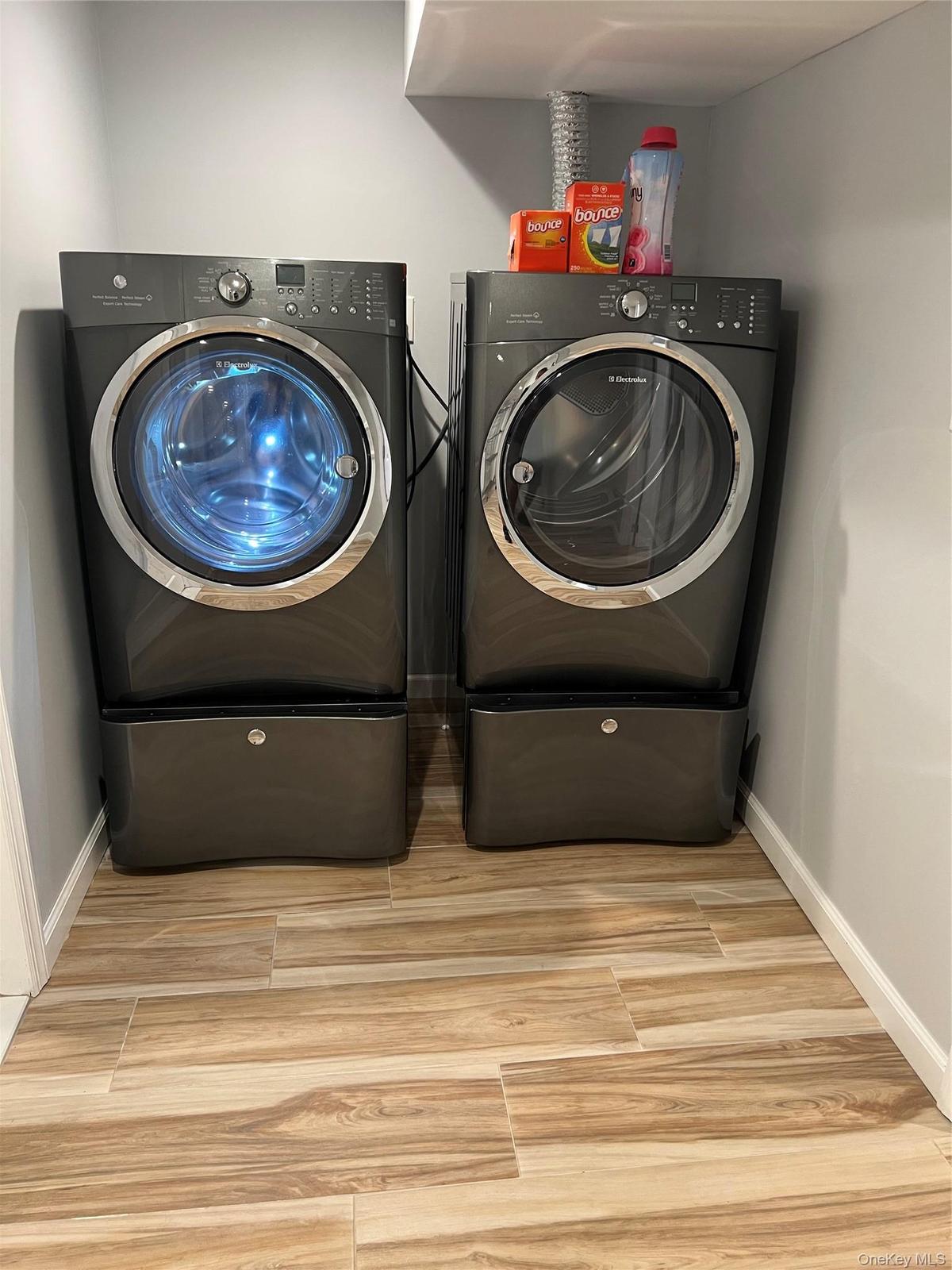 Dedicated laundry room featuring side-by-side front-load washers and dryers with pedestal bases, ample countertop storage above, wood-finish flooring,