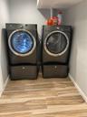 Dedicated laundry room featuring side-by-side front-load washers and dryers with pedestal bases, ample countertop storage above, wood-finish flooring,