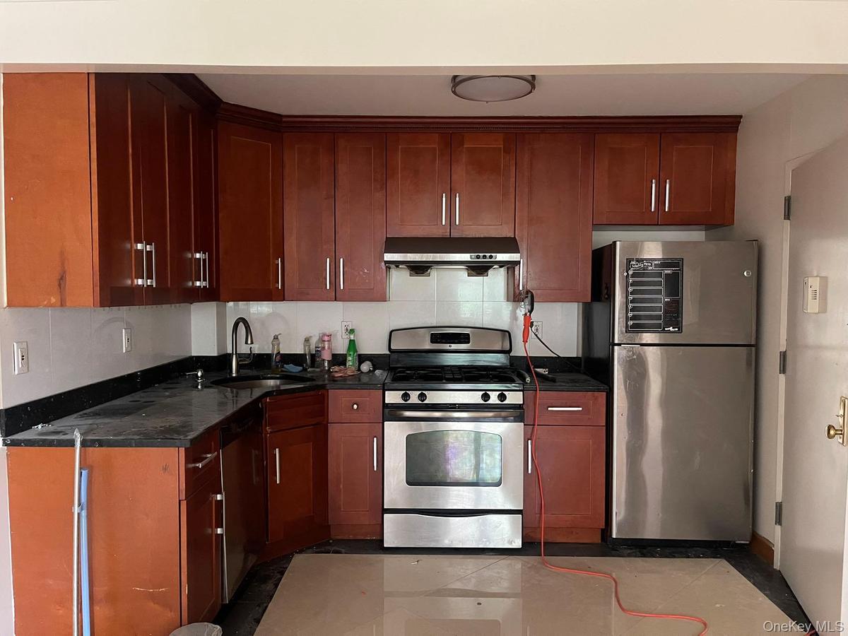 Kitchen with stainless steel appliances, backsplash, range hood, light tile patterned flooring, and dark stone counters